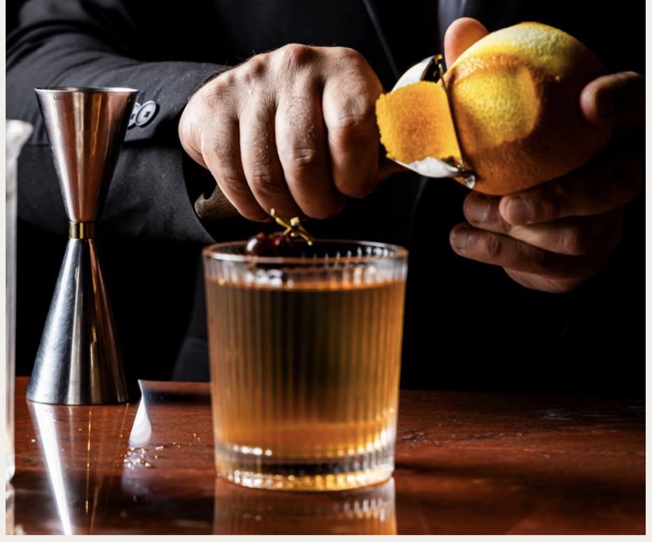 Mãos retirando a casca de uma laranja sobre um coquetel no balcão do bar